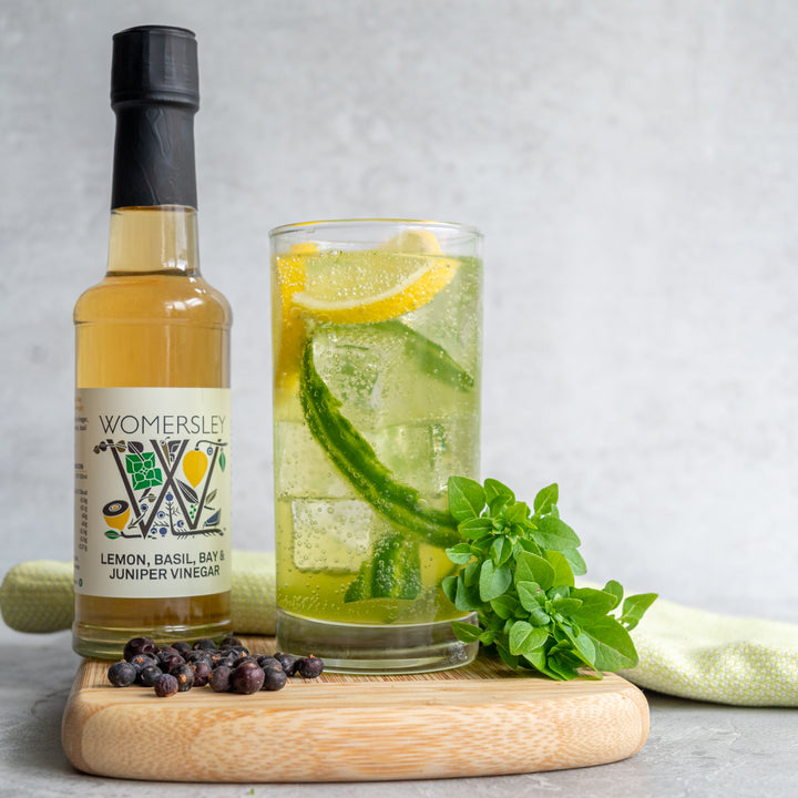 Womersley Foods 150ml Lemon, Basil, Bay & Juniper Fruit Vinegar standing on a cutting board with a glass of ice-cold lemon and basil water sounded by other ingredients and a napkin.