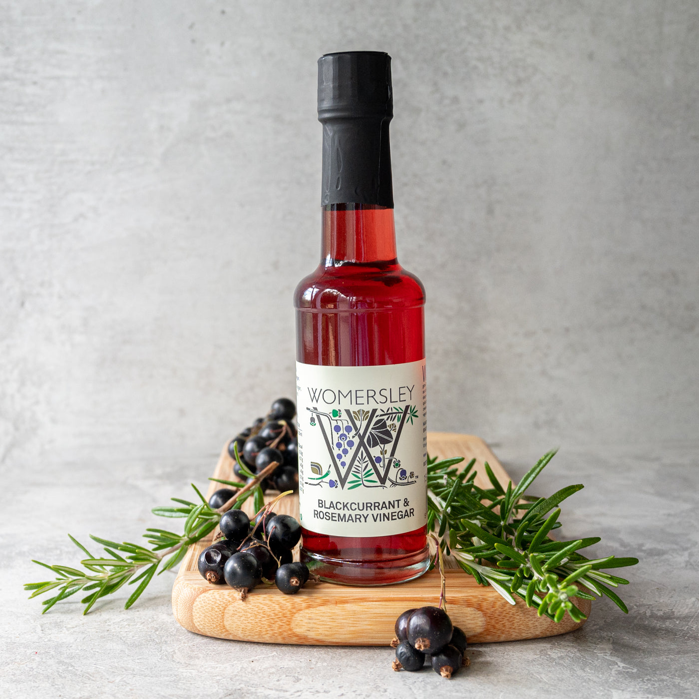 Womersley Foods Blackcurrant & Rosemary Fruit Vinegar standing on a cutting board with grey background surrounded by blackcurrant and rosemary ingredients.