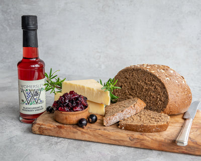 Womersley Foods Blackcurrant & Rosemary Fruit Vinegar standing on a cutting board with grey background surrounded by blackcurrant and rosemary ingredients and slices of bread and blackcurrant jam and cheese on a cutting board with knife.