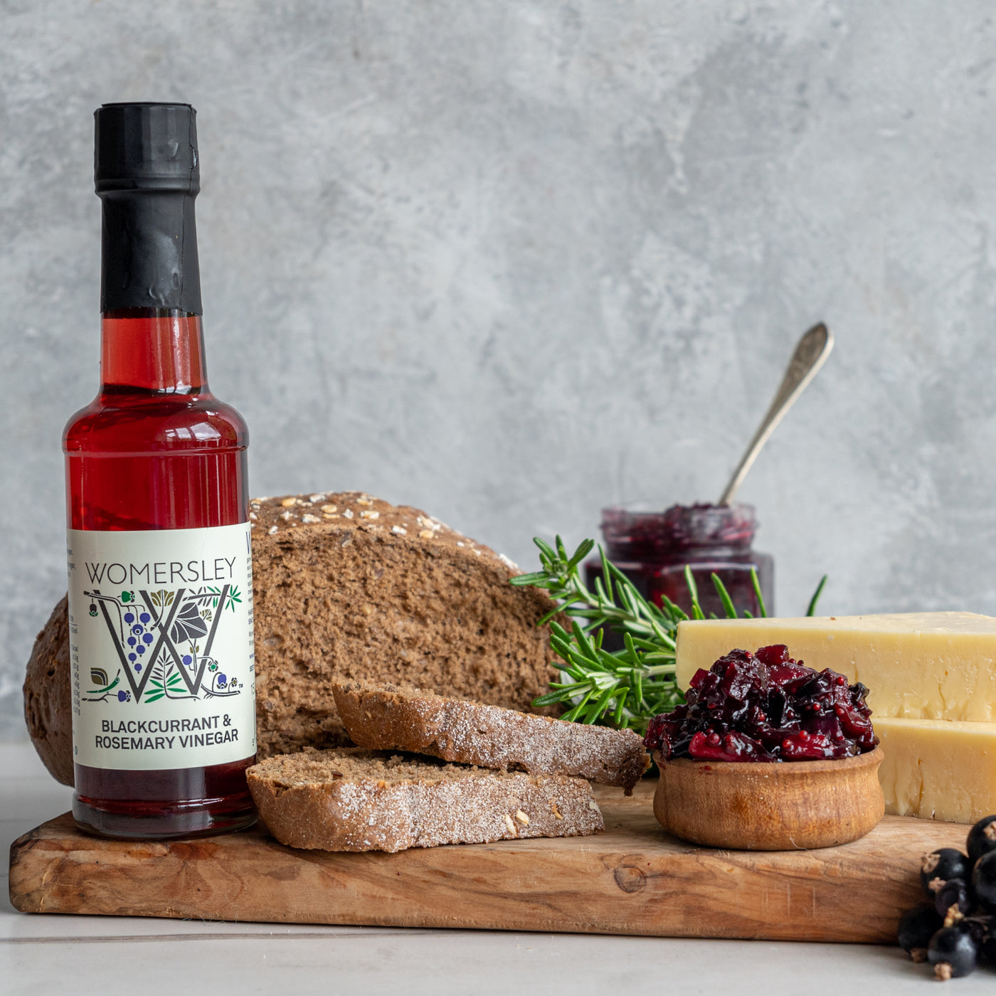 Womersley Foods Blackcurrant & Rosemary Fruit Vinegar standing on a cutting board with grey background surrounded by blackcurrant and rosemary ingredients and slices of bread and blackcurrant jam and cheese on a cutting board.