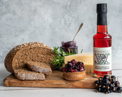 Womersley Foods Blackcurrant & Rosemary Fruit Vinegar on the right side standing on a cutting board with grey background surrounded by blackcurrant and rosemary ingredients and slices of bread and blackcurrant jam and cheese on a cutting board.