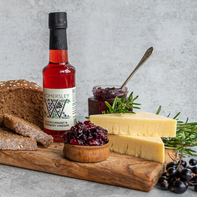 Clsoe up Womersley Foods Blackcurrant & Rosemary Fruit Vinegar on the left side standing on a cutting board with grey background surrounded by blackcurrant and rosemary ingredients and slices of bread and blackcurrant jam and cheese on a cutting board.