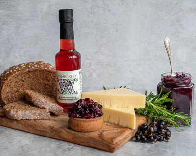 Womersley Foods Blackcurrant & Rosemary Fruit Vinegar on the left side standing on a cutting board with grey background surrounded by blackcurrant and rosemary ingredients and slices of bread and blackcurrant jam and cheese on a cutting board.
