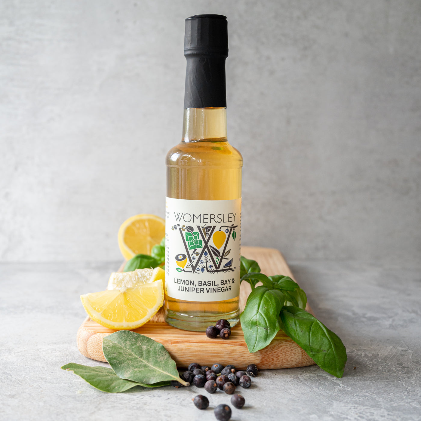 Womersley Foods 150ml Lemon, Basil, Bay & Juniper Fruit Vinegar standing on a cutting board with grey background surrounded by lemon, basil, bay and juniper ingredients.