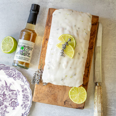 Top view of Lime, Black Pepper and Lavender Drizzle cake on a cutting board made with Womersley Foods Lime, Black Pepper & Lavender Fruit Vinegar. Surrounded by ingredients, a bottle of Womersley Foods 150ml Lime, Black Pepper & Lavender Fruit Vinegar and a bread knife and plate.