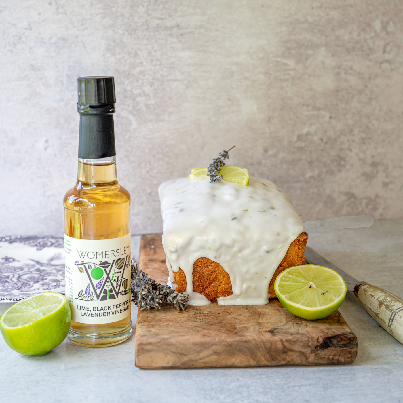 Front view of Lime, Black Pepper and Lavender Drizzle cake on a cutting board made with Womersley Foods Lime, Black Pepper & Lavender Fruit Vinegar. Surrounded by ingredients, a bottle of Womersley Foods 150ml Lime, Black Pepper & Lavender Fruit Vinegar and a bread knife and plate.