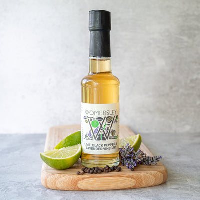 Womersley Foods 150ml Lime, Black Pepper & Lavender Fruit Vinegar standing on a cutting board with grey background surrounded by Lime, Black Pepper & Lavender ingredients on a grey background.