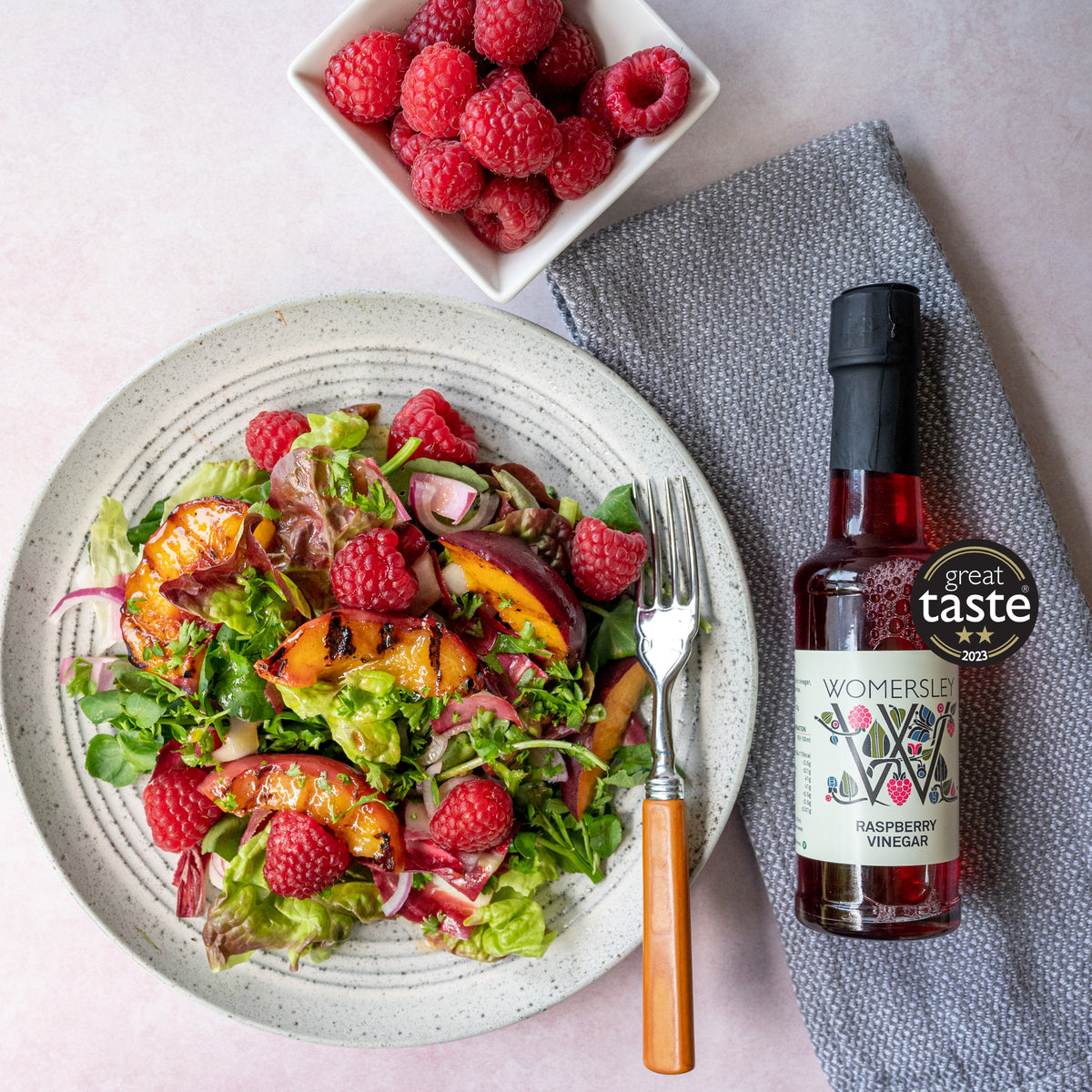 Sqaure image of a bottle of Womersley Foods Raspberry Fruit Vinegar used on a plate of delicious Raspberry and lettuce salad with slices of bread and a bowl of Raspberry with a fork and napkin.
