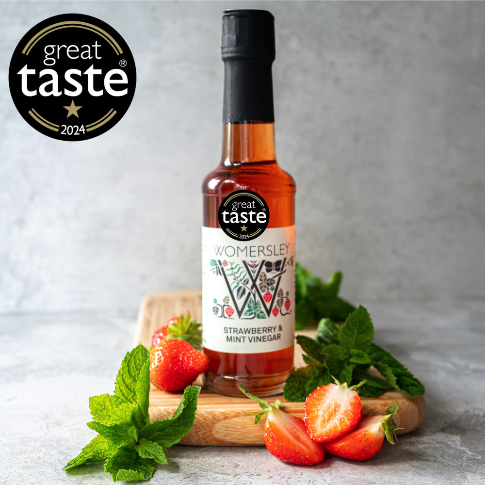 A bottle of Strawberry & Mint Fruit & Herb Vinegar on a wooden board, surrounded by fresh strawberries and mint leaves. There's a 'Great Taste 2021' award badge displayed in the top left corner.