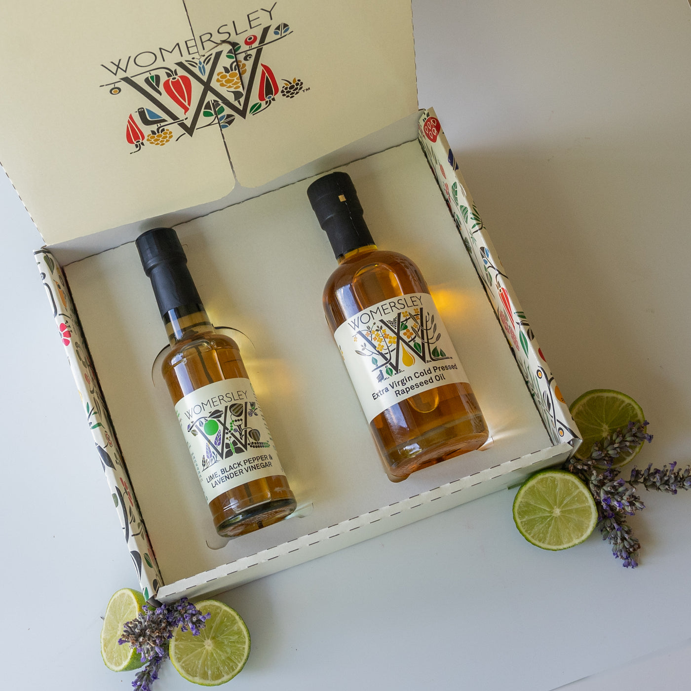 Womersley Foods Fruit & Herb vinegar opened Gift Box Divinely Luxurious Dressing with ingredients. Featuring lime, black pepper and lavender fruit vinegar and extra virgin cold pressed rapeseed oil with image of cut lime and lavender on a grey background.