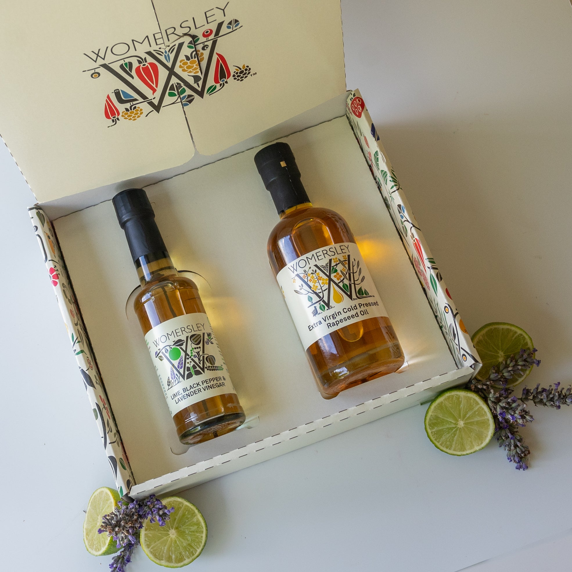 Womersley Foods Fruit & Herb vinegar closed Gift Box Divinely Luxurious Dressing with ingredients. Featuring lime, black pepper and lavender fruit vinegar and extra virgin cold pressed rapeseed oil standing in front of box with image of cut lime and lavender on a grey background.