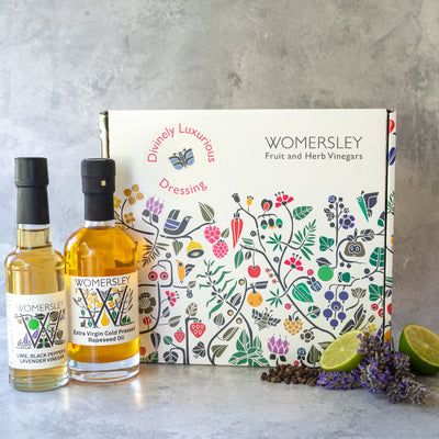 Womersley Foods Fruit & Herb vinegar closed Gift Box Divinely Luxurious Dressing with ingredients. Featuring lime, black pepper and lavender fruit vinegar and extra virgin cold pressed rapeseed oil standing in front of box with image of cut lime and lavender on a grey background.