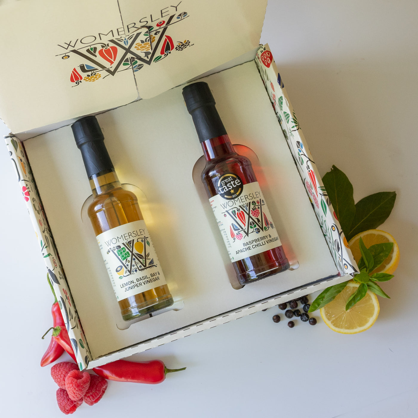 Womersley Foods Fruit & Herb vinegar opened Gift Box Enduringly Popular Vinegars with ingredients. Featuring 150ml Raspberry and Apache Chili fruit vinegar and Lemon, basil, Bay and Juniper fruit vinegar lying in the box with images of raspberry, lemon, basil, bay and Apache chili on a white background.