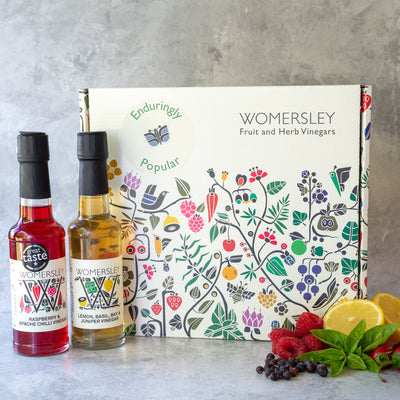 Womersley Foods Fruit & Herb vinegar closed Gift Box Enduringly Popular Vinegars with ingredients. Featuring 150ml Raspberry and Apache Chili fruit vinegar and Lemon, basil, Bay and Juniper fruit vinegar standing in front of box with image of raspberry, lemon, basil, bay and Apache chili on a grey background.