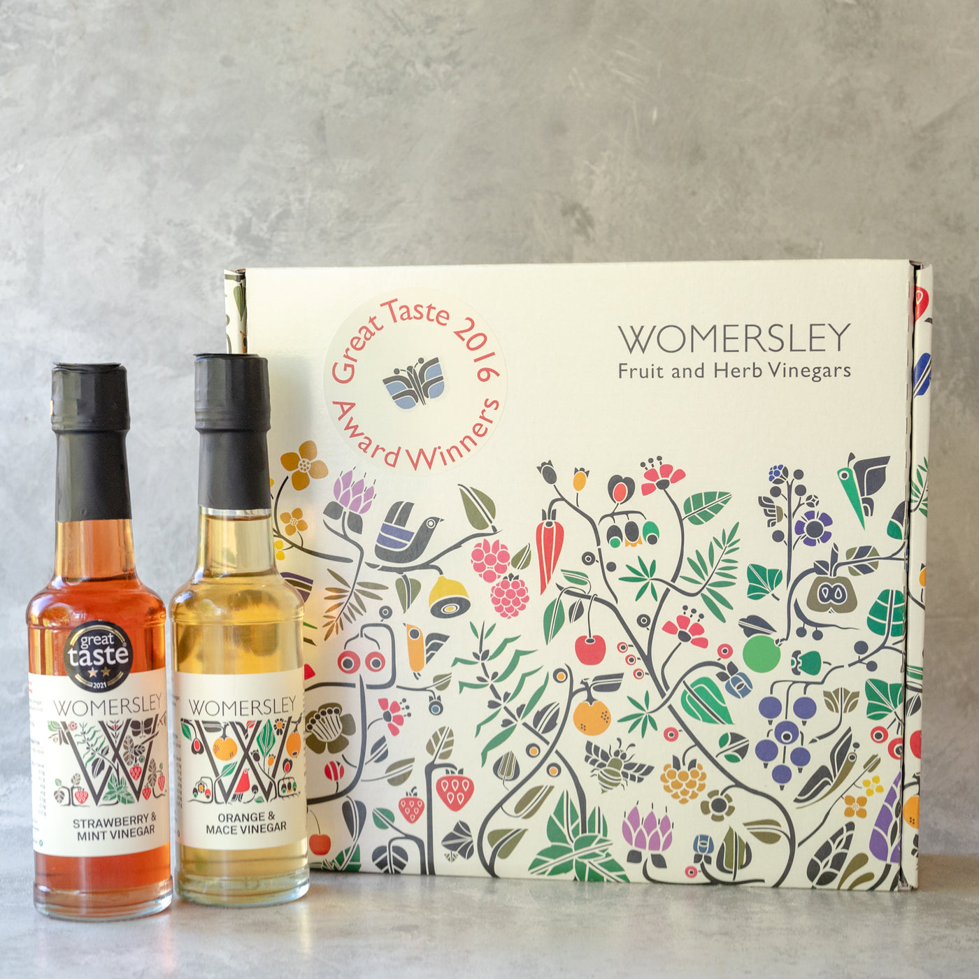 Womersley Foods Fruit & Herb vinegar closed Gift Box Great Taste Award Vinegars, with ingredients. Featuring 150ml Strawberry and Mint fruit vinegar and Orange and Mace fruit vinegar standing in front of box on a grey background.