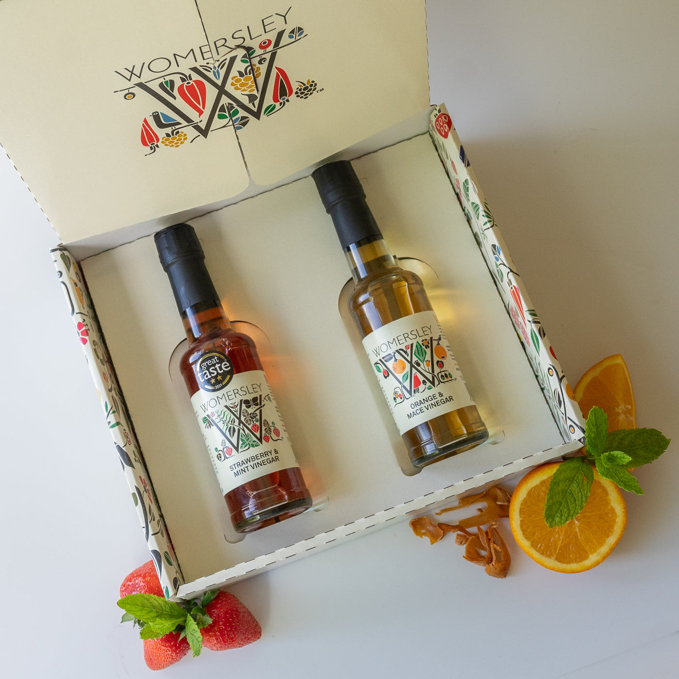 Womersley Foods Fruit & Herb vinegar open Gift Box Great Taste Award Vinegars, with ingredients. Featuring 150ml Strawberry and Mint fruit vinegar and Orange and Mace fruit vinegar lying in the box with image of strawberry, mint leaf, orange and mace on a white background.