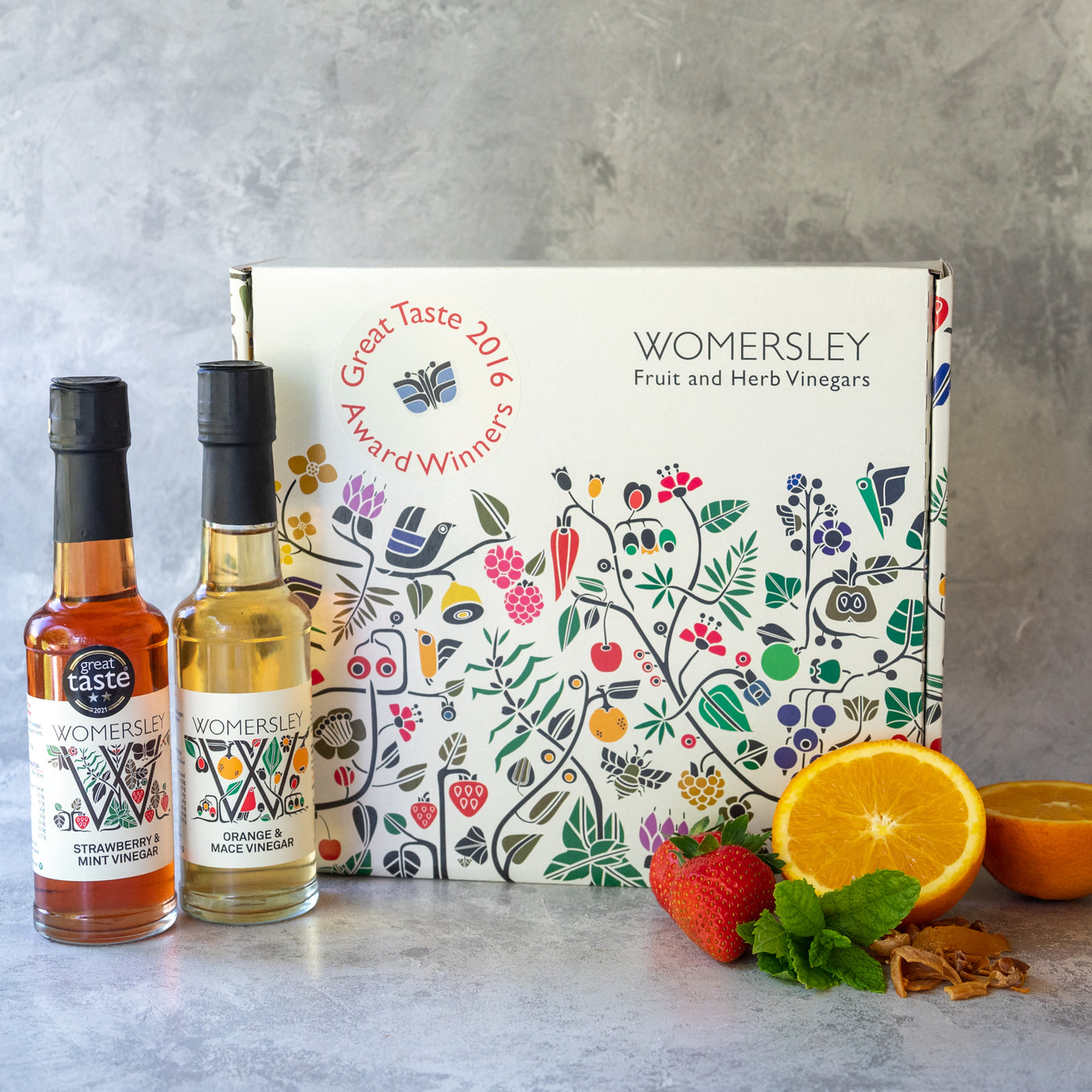Womersley Foods Fruit & Herb vinegar closed Gift Box Great Taste Award Vinegars, with ingredients. Featuring 150ml Strawberry and Mint fruit vinegar and Orange and Mace fruit vinegar standing in front of box with image of strawberry, mint leaf, orange and mace on a grey background.