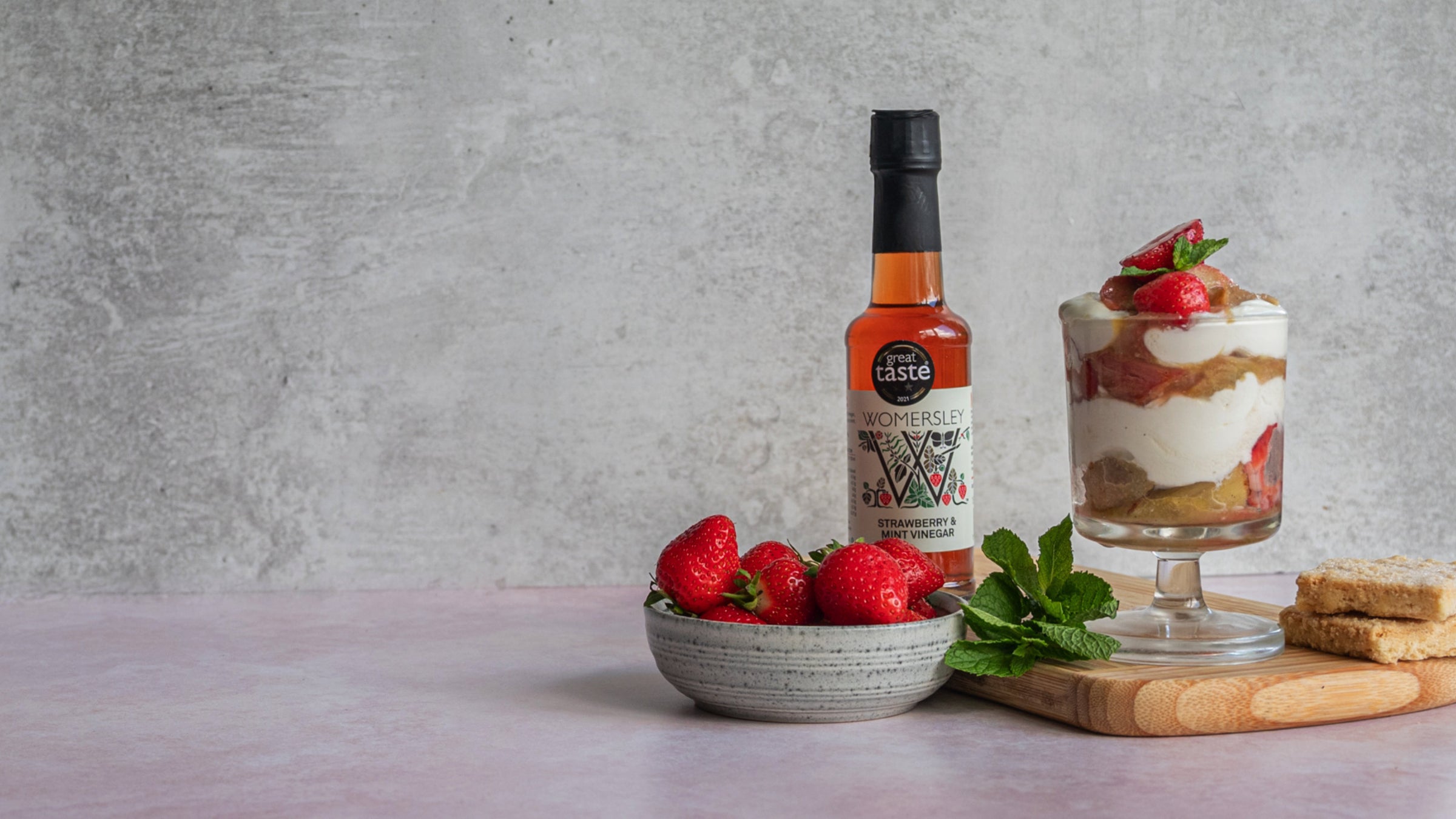 Womersley Foods Strawberry & Mint Vinegar bottle used to make strawberry ice cream in an ice cream cup with a bowl of strawberry, mint leaf and bread on a cutting board.