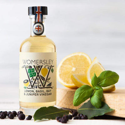 Womersley Foods 100ml Lemon, Basil, Bay & Juniper Fruit Vinegar standing beside a cutting board surrounded by lemon, basil, bay and juniper ingredients.